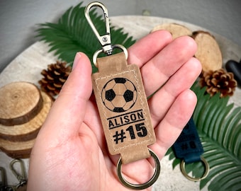 May include: Brown leather keychain with a gold metal ring and a black soccer ball design. The keychain is engraved with the name "Alison" and the number "#15".