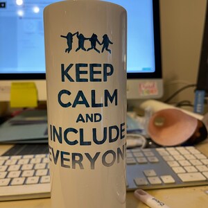 May include: White tumbler with the text "KEEP CALM AND INCLUDE EVERYONE" in blue. Above the text are four blue silhouettes of people holding hands and jumping. The tumbler is cylindrical and appears to be made of ceramic or a similar material.
