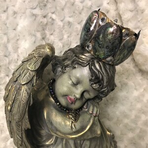 May include: A sleeping angel figurine with wings, a crown made of iridescent abalone shell, and a heart-shaped pendant necklace. The angel is painted in shades of green and gold.