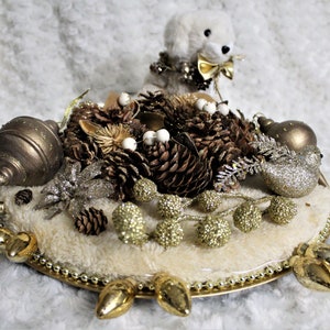 Puede incluir: Una base de piel sintética blanca con un borde de metal dorado y detalles dorados. La base está decorada con piñas, adornos dorados y un animal de peluche blanco.