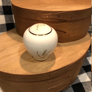 Peut inclure: Une salière en céramique blanche de forme arrondie, dotée d'un bord doré sur le dessus et d'un motif de tige de blé dorée sur le côté. La salière possède un couvercle perforé avec de petits trous pour le sel et repose sur une surface lisse.
