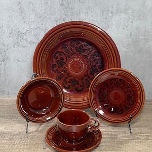 May include: A set of brown ceramic dinnerware featuring a large plate, two bowls, and a cup with a saucer. Each piece displays a dark brown embossed floral pattern against a glossy brown glaze, with concentric ring details along the outer rims.