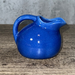 May include: A small, round ceramic creamer jug with a vibrant cobalt blue glaze. The jug features a curved handle on the left side and a distinct pouring spout on the right, with a smooth, glossy finish reflecting light across its rounded body.