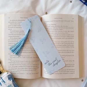 Puede incluir: Marcapáginas azul claro con borla, con un diseño de palomas y la frase "onto the next chapter". Se encuentra en un libro abierto, perfecto para la lectura.