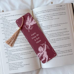 May include: Burgundy bookmark with a tassel, featuring a Cupid design with a bow and arrow, and the text "Cupid, Bring Me A Hot Villain." The bookmark is placed on an open book.