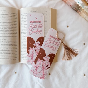 May include: Two rectangular bookmarks with the text "WEAR THE HAT Ride the Cowboy" in pink script. The bookmarks feature a pink illustration of a cowgirl on a horse. A book is open in the background.