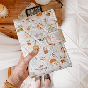 Può includere: Una custodia per libri in tessuto trapuntato con sfondo bianco e un motivo ripetuto di piccole illustrazioni che includono uccelli, piante in vaso, gomitoli di lana, libri e case. La custodia ha una chiusura a bottone automatico e un'etichetta JOLI STUDIO.
