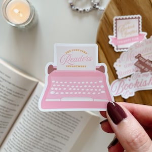 Op de afbeelding: Roze typemachine sticker met de tekst "The Tortured Readers Department" in gouden letters.