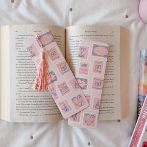 May include: Two pink bookmarks with a stamp design featuring hearts, wedding rings, and the word "DAD". One bookmark has a pink tassel. The bookmarks are placed on an open book.