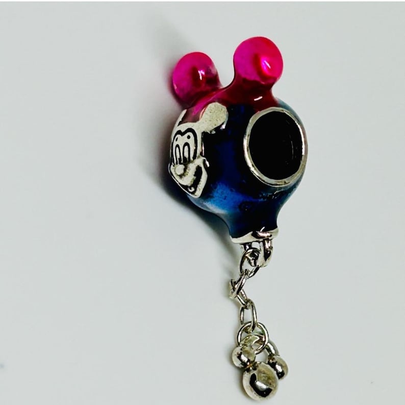 Puede incluir: Un charm con un dise&ntilde;o de rat&oacute;n de dibujos animados. El charm tiene un cuerpo azul y plateado con una cabeza y orejas rosas. Una cadena plateada con tres peque&ntilde;as cuentas plateadas cuelga de la parte inferior.