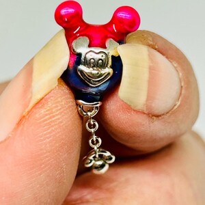 Puede incluir: Un charm de Mickey Mouse con un dise&ntilde;o rojo y azul. El charm tiene una cara plateada y un acento floral colgante. El charm se sostiene entre dos dedos.