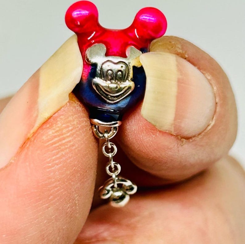 Puede incluir: Un charm de Mickey Mouse con un dise&ntilde;o rojo y azul. El charm tiene una cara plateada y un acento floral colgante. El charm se sostiene entre dos dedos.
