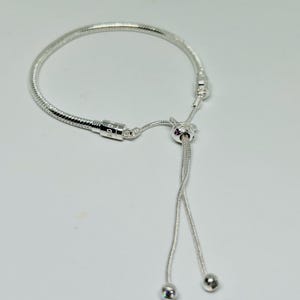 K&ouml;nnte beinhalten: Ein silbernes Armband mit Schlangenketten-Design und einem Schiebeverschluss. Das Armband hat eine kleine silberne Perle am Ende der Schiebeschnur.