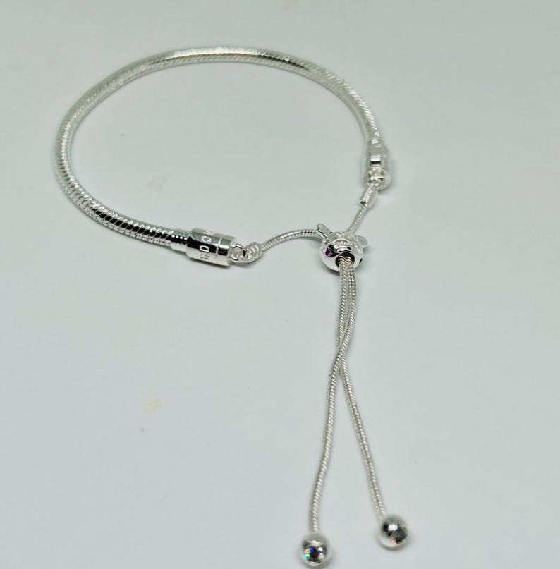 K&ouml;nnte beinhalten: Ein silbernes Armband mit Schlangenketten-Design und einem Schiebeverschluss. Das Armband hat eine kleine silberne Perle am Ende der Schiebeschnur.