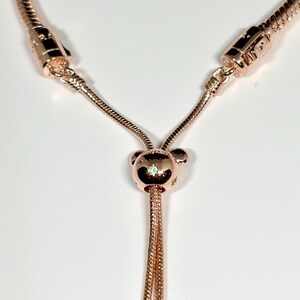 Op de afbeelding: Ros&eacute;goudkleurige ketting met een schuifsluiting en een centrale bedel. De bedel heeft een beerachtig ontwerp met kleine, kleurrijke accenten. De ketting heeft een slangenkettingontwerp en wordt gepresenteerd op een witte achtergrond.