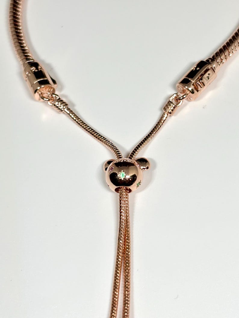 Op de afbeelding: Ros&eacute;goudkleurige ketting met een schuifsluiting en een centrale bedel. De bedel heeft een beerachtig ontwerp met kleine, kleurrijke accenten. De ketting heeft een slangenkettingontwerp en wordt gepresenteerd op een witte achtergrond.