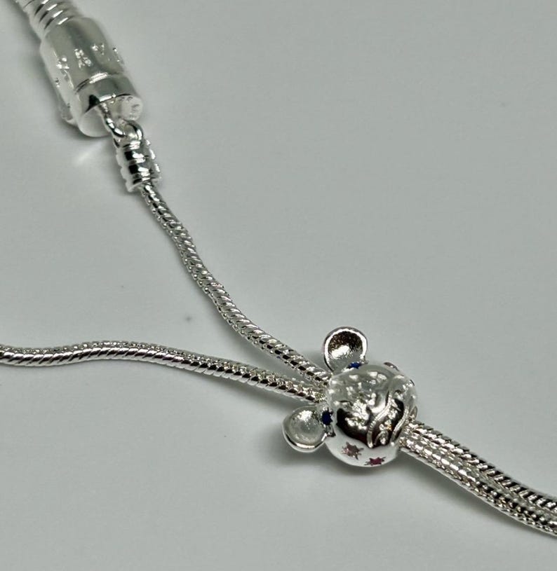 K&ouml;nnte beinhalten: Ein silbernes Kettenarmband mit einem silbernen Anh&auml;nger in Form eines Comic-Mauskopfes mit blauen und roten Edelsteinen.