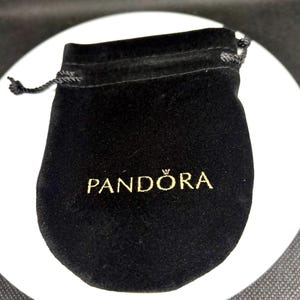Op de afbeelding: Een zwarte fluwelen zakje met een trekkoordsluiting. Op de voorkant van het zakje staat het woord "PANDORA" in gouden letters gedrukt. Het zakje wordt waarschijnlijk gebruikt voor het opbergen van sieraden of kleine items.