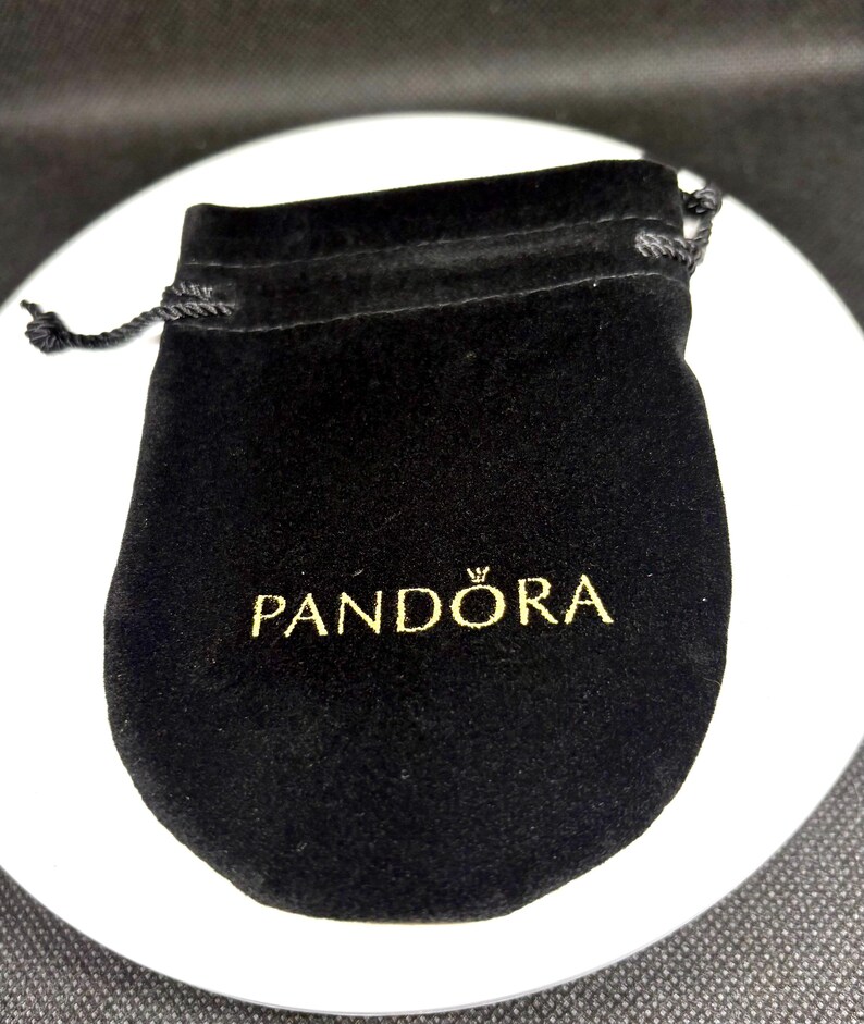 Op de afbeelding: Een zwarte fluwelen zakje met een trekkoordsluiting. Op de voorkant van het zakje staat het woord "PANDORA" in gouden letters gedrukt. Het zakje wordt waarschijnlijk gebruikt voor het opbergen van sieraden of kleine items.