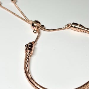 Op de afbeelding: Ros&eacute;gouden armband met een slangenkettingontwerp. De armband heeft een schuifsluiting en een decoratieve kraal met kleine, kleurrijke accenten. Het metaal heeft een gepolijste afwerking, die licht reflecteert. De armband wordt tegen een witte achtergrond getoond.