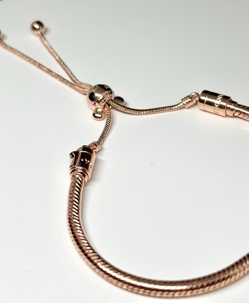 Op de afbeelding: Ros&eacute;gouden armband met een slangenkettingontwerp. De armband heeft een schuifsluiting en een decoratieve kraal met kleine, kleurrijke accenten. Het metaal heeft een gepolijste afwerking, die licht reflecteert. De armband wordt tegen een witte achtergrond getoond.