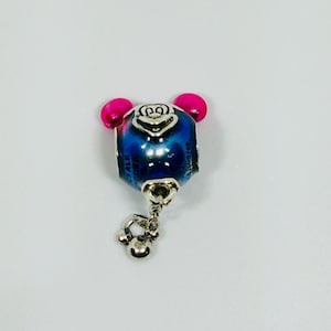 Puede incluir: Un charm azul y plateado con detalles en rosa. El charm tiene un dise&ntilde;o similar a un oso con un charm plateado colgante. El charm probablemente est&aacute; hecho de metal y est&aacute; dise&ntilde;ado para agregarse a una pulsera o collar.