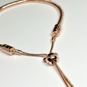 Op de afbeelding: Ros&eacute;goudkleurige armband met een slangenkettingontwerp. De armband heeft een schuifsluiting en een decoratieve kraal met stervormige accenten en een kleine groene edelsteen. De armband wordt getoond tegen een witte achtergrond.