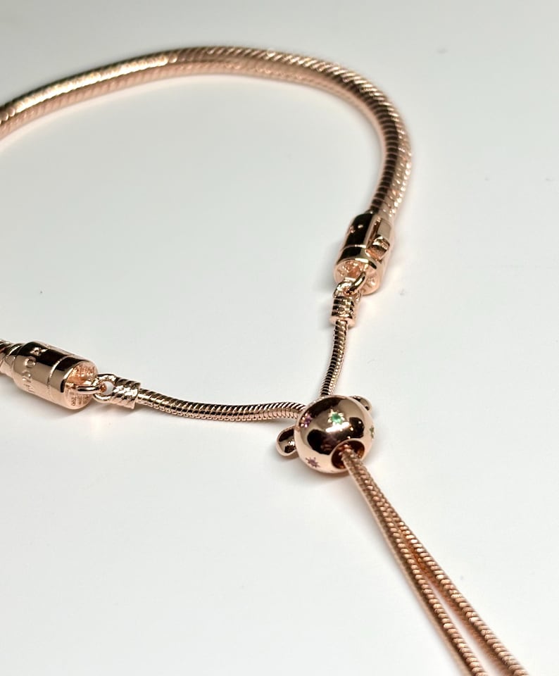 Op de afbeelding: Ros&eacute;goudkleurige armband met een slangenkettingontwerp. De armband heeft een schuifsluiting en een decoratieve kraal met stervormige accenten en een kleine groene edelsteen. De armband wordt getoond tegen een witte achtergrond.