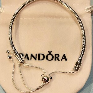 K&ouml;nnte beinhalten: Ein silbernes Pandora-Armband mit einem Schlangenketten-Design und einem Schiebeverschluss. Das Armband hat einen kleinen, runden Anh&auml;nger mit einem rosa Stein am Ende des Schiebeverschlusses.