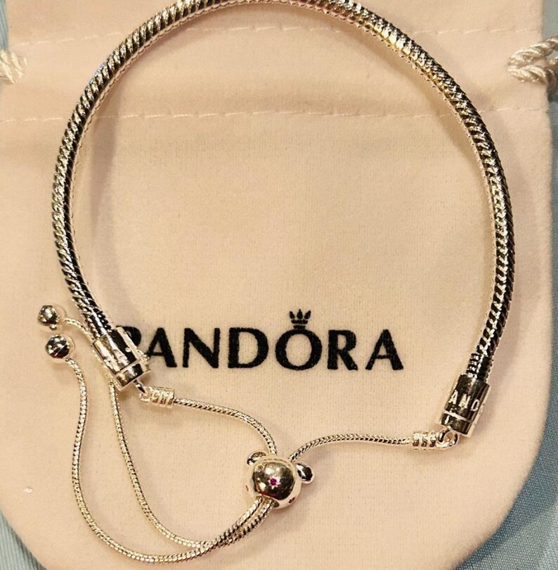 K&ouml;nnte beinhalten: Ein silbernes Pandora-Armband mit einem Schlangenketten-Design und einem Schiebeverschluss. Das Armband hat einen kleinen, runden Anh&auml;nger mit einem rosa Stein am Ende des Schiebeverschlusses.