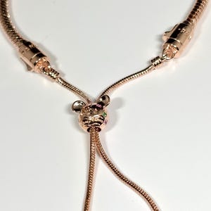 Op de afbeelding: Verstelbare armband in ros&eacute;goud met een decoratieve bedel. De bedel heeft een kleine dierenkop met kleurrijke edelstenen. De armband heeft een slangenketen ontwerp en een schuifsluiting.