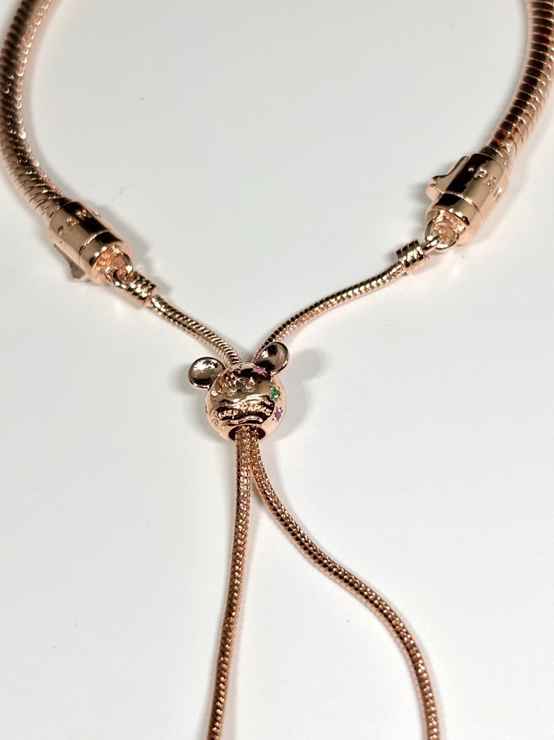 Op de afbeelding: Verstelbare armband in ros&eacute;goud met een decoratieve bedel. De bedel heeft een kleine dierenkop met kleurrijke edelstenen. De armband heeft een slangenketen ontwerp en een schuifsluiting.