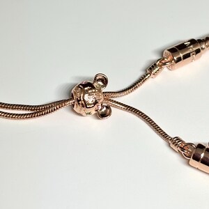 Op de afbeelding: Ros&eacute;gouden armband met een Mickey Mouse bedel. De bedel heeft kleine groene details. De armband heeft een slangenketting ontwerp en een veilige sluiting. De sieraden worden gepresenteerd tegen een witte achtergrond.