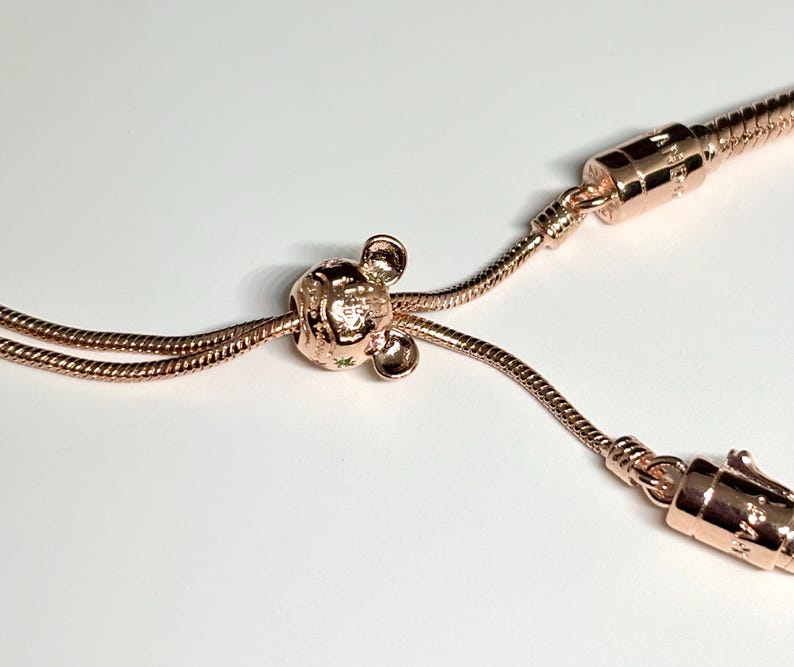 Op de afbeelding: Ros&eacute;gouden armband met een Mickey Mouse bedel. De bedel heeft kleine groene details. De armband heeft een slangenketting ontwerp en een veilige sluiting. De sieraden worden gepresenteerd tegen een witte achtergrond.