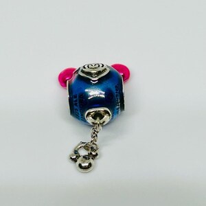 Puede incluir: Un charm azul y plateado con detalles en rosa. El charm tiene un dise&ntilde;o de Mickey Mouse y se ven las palabras "Disney" y "925 ALE Pandora". Un peque&ntilde;o charm plateado cuelga de la parte inferior.