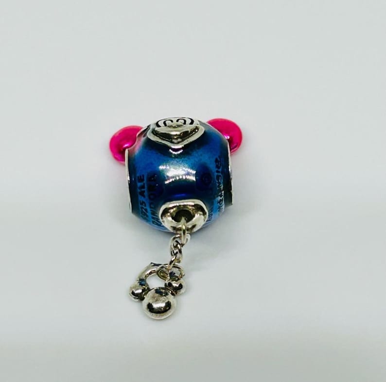 Puede incluir: Un charm azul y plateado con detalles en rosa. El charm tiene un dise&ntilde;o de Mickey Mouse y se ven las palabras "Disney" y "925 ALE Pandora". Un peque&ntilde;o charm plateado cuelga de la parte inferior.