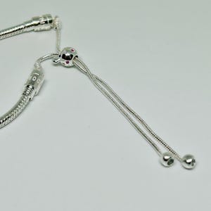 K&ouml;nnte beinhalten: Ein silbernes Schlangenkettenarmband mit einem silbernen Perlenanh&auml;nger und einem silbernen Schiebeverschluss. Der Perlenanh&auml;nger hat kleine rosa Steine.