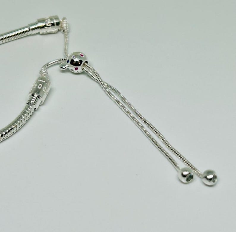 K&ouml;nnte beinhalten: Ein silbernes Schlangenkettenarmband mit einem silbernen Perlenanh&auml;nger und einem silbernen Schiebeverschluss. Der Perlenanh&auml;nger hat kleine rosa Steine.