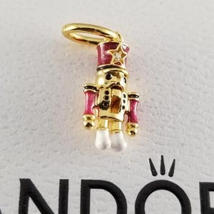 May include: A gold-colored charm in the shape of a toy soldier, featuring a red and gold hat with a star, red accents, and white boots. The charm has a loop for attaching to a bracelet or necklace.