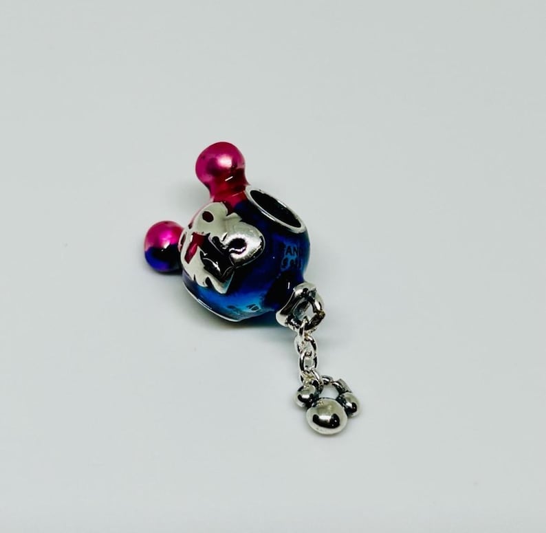 Puede incluir: Un charm con un degradado azul y morado, con un dise&ntilde;o plateado y un charm colgante de huella de pata. El charm tiene una parte superior rosa y una peque&ntilde;a esfera rosa. El charm est&aacute; sobre un fondo blanco.