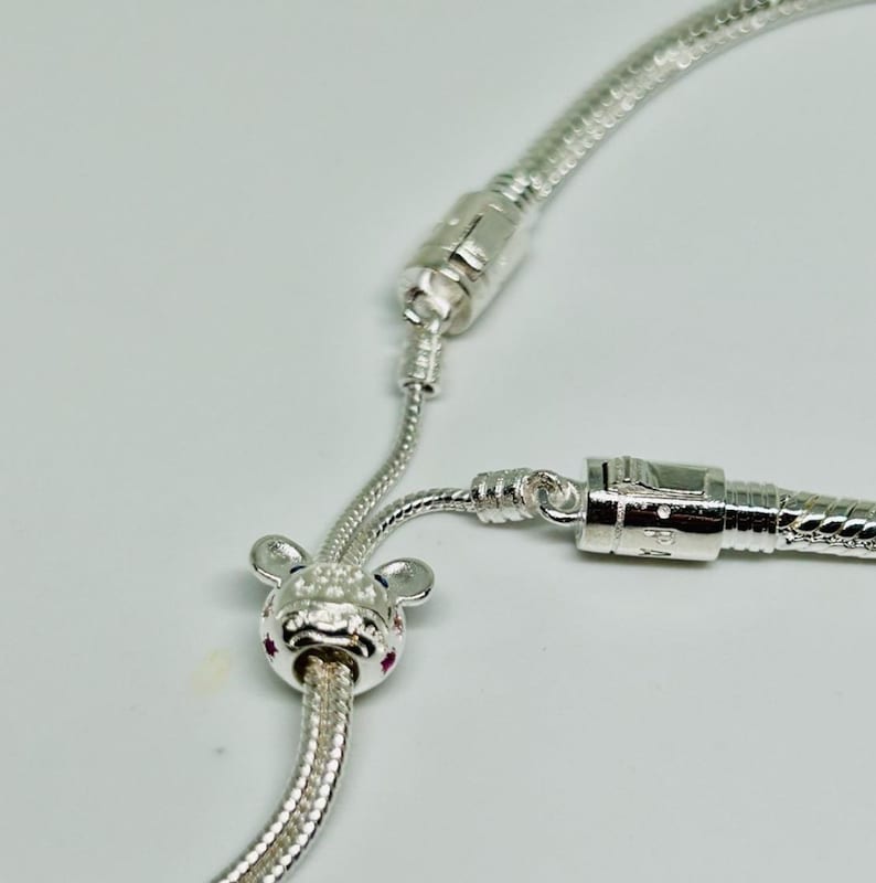 K&ouml;nnte beinhalten: Ein silbernes Armband mit einem silbernen Anh&auml;nger in Form einer Comic-Maus mit rosa und blauen Akzenten. Das Armband hat einen silbernen Verschluss und eine silberne Verl&auml;ngerungskette.