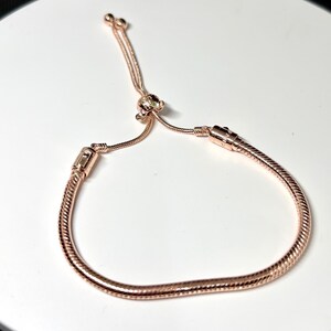 Op de afbeelding: Verstelbare armband in ros&eacute;goud met een slangenkettingontwerp. De armband heeft een schuifsluiting en een kleine decoratieve bedel. Het metaal heeft een glanzende afwerking, die licht reflecteert.