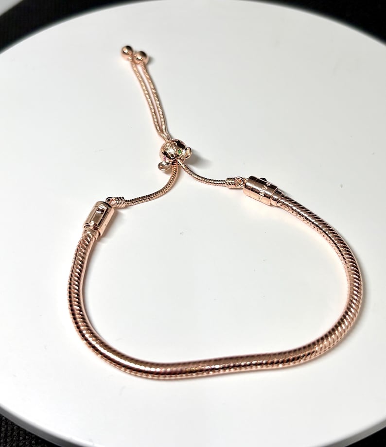 Op de afbeelding: Verstelbare armband in ros&eacute;goud met een slangenkettingontwerp. De armband heeft een schuifsluiting en een kleine decoratieve bedel. Het metaal heeft een glanzende afwerking, die licht reflecteert.