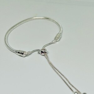 K&ouml;nnte beinhalten: Ein silbernes Armband mit einem Schiebeperlenverschluss und einem kleinen Anh&auml;nger. Das Armband ist aus einer Schlangenkettenform gefertigt.