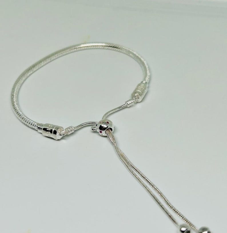 K&ouml;nnte beinhalten: Ein silbernes Armband mit einem Schiebeperlenverschluss und einem kleinen Anh&auml;nger. Das Armband ist aus einer Schlangenkettenform gefertigt.