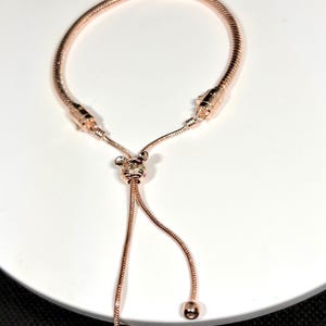 Op de afbeelding: Ros&eacute;gouden verstelbare armband met een slangenkettingontwerp. De armband heeft een schuifsluiting en decoratieve accenten. Het verstelbare ontwerp zorgt voor een aangepaste pasvorm. De armband wordt gepresenteerd op een wit oppervlak.