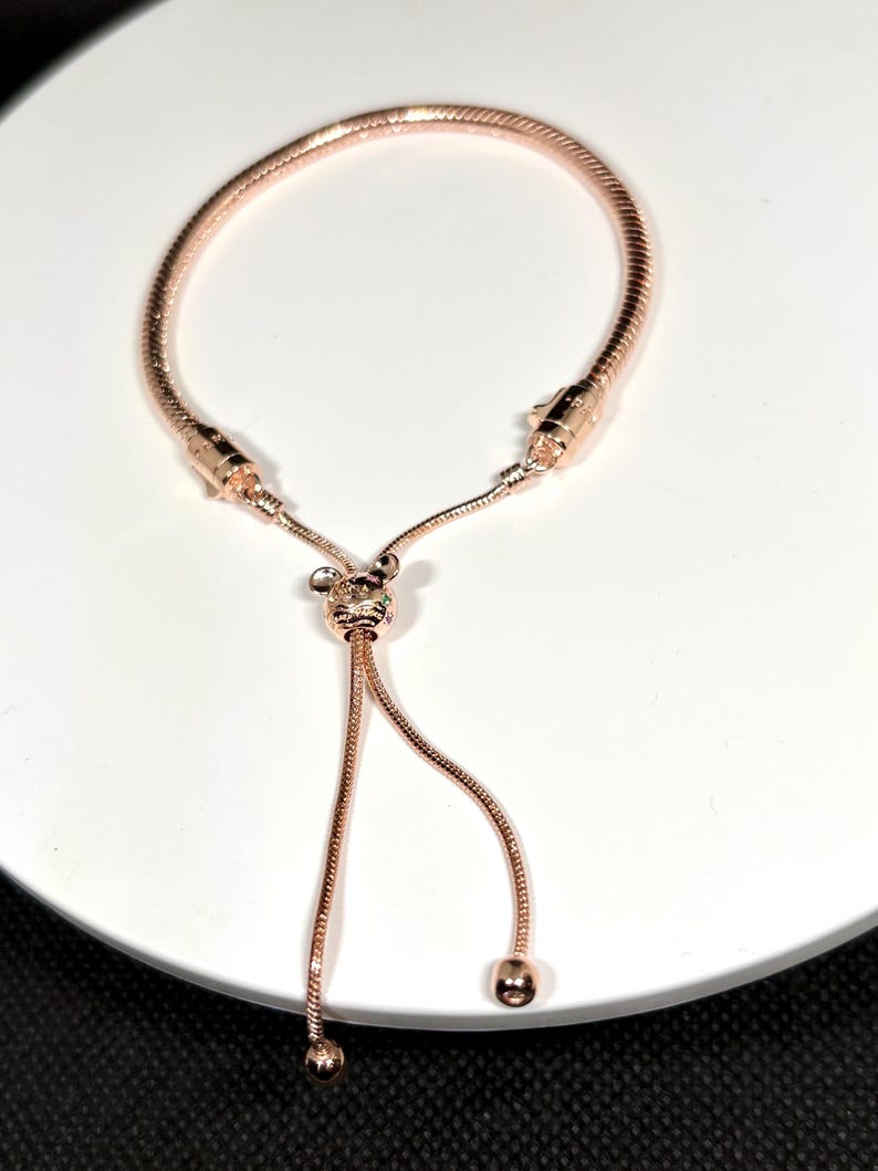 Op de afbeelding: Ros&eacute;gouden verstelbare armband met een slangenkettingontwerp. De armband heeft een schuifsluiting en decoratieve accenten. Het verstelbare ontwerp zorgt voor een aangepaste pasvorm. De armband wordt gepresenteerd op een wit oppervlak.