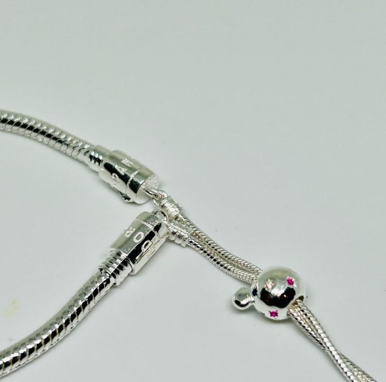 K&ouml;nnte beinhalten: Ein silbernes Armband mit einem kleinen, runden Anh&auml;nger mit rosa Steinen. Das Armband hat einen Schiebeverschluss und ist aus einer Schlangenkette gefertigt.