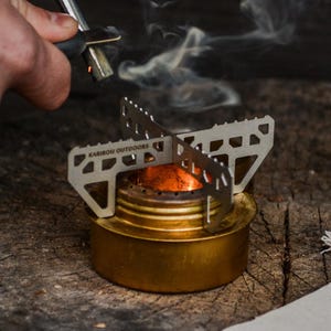 Op de afbeelding: Een goudkleurige metalen campingbrander met een zilveren kruisvormige pannendrager, een donker olijfgroene Primus waterfles en een klein gebroken wit stoffen trekkoordzakje met een hertlogo en de tekst Karibou Outdoors.