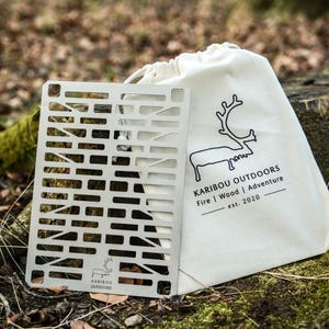 Puede incluir: Una parrilla de metal y una bolsa de lona con el texto "KARIBOU OUTDOORS" y un gráfico de reno. La parrilla tiene forma rectangular con un patrón de formas recortadas. La bolsa es blanca con cierre de cordón.